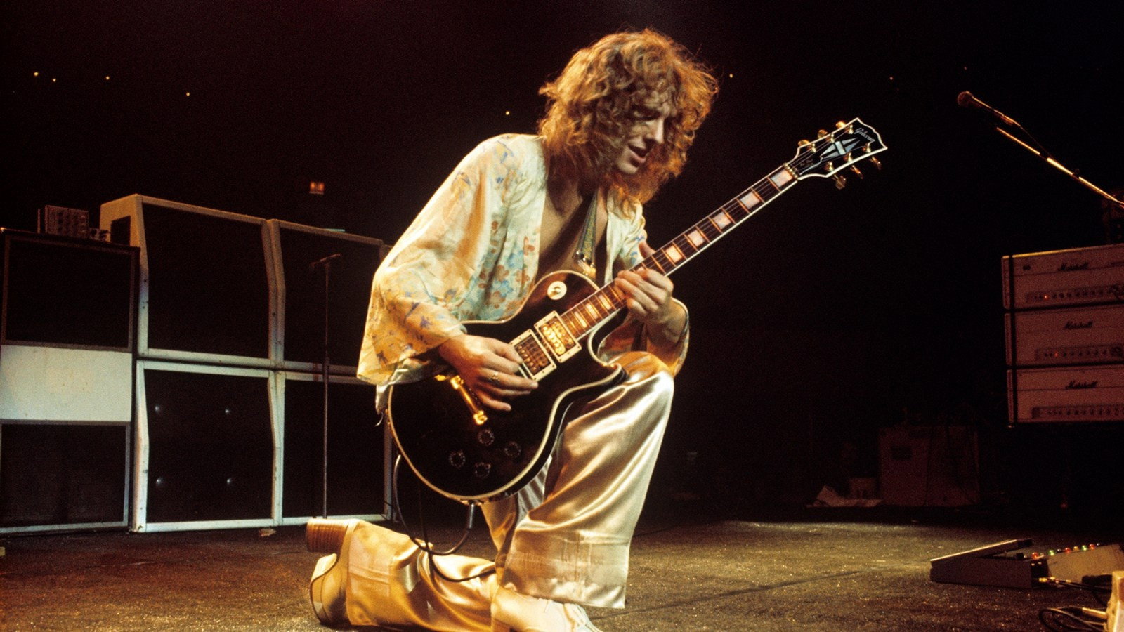 A young Frampton in a satin outfit kneeling on stage with a black Les Paul, Marshall amplifiers behind him