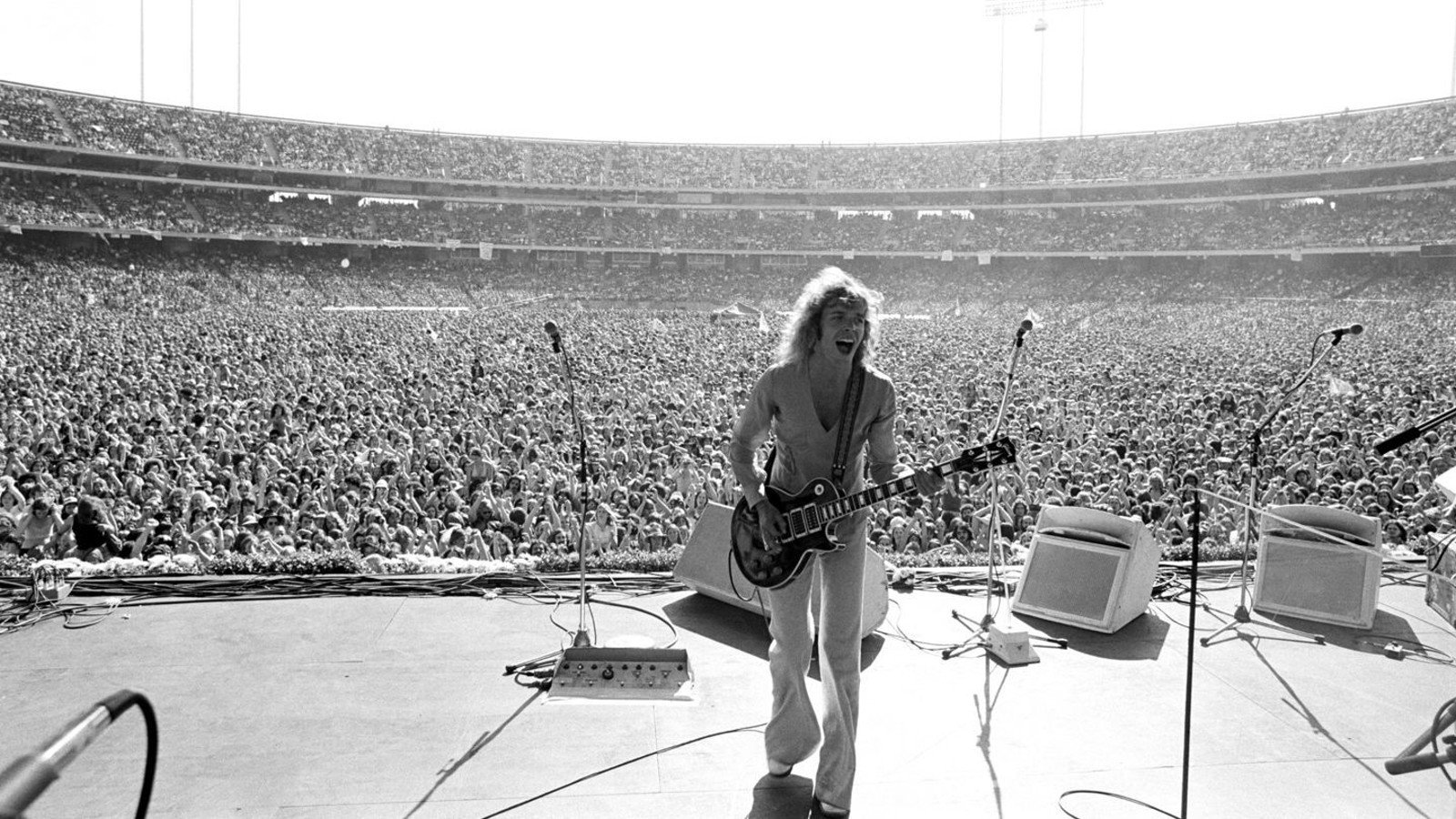 Black-and-white photograph: Frampton on a stadium stage with his guitar, a crowd filling the stands behind him