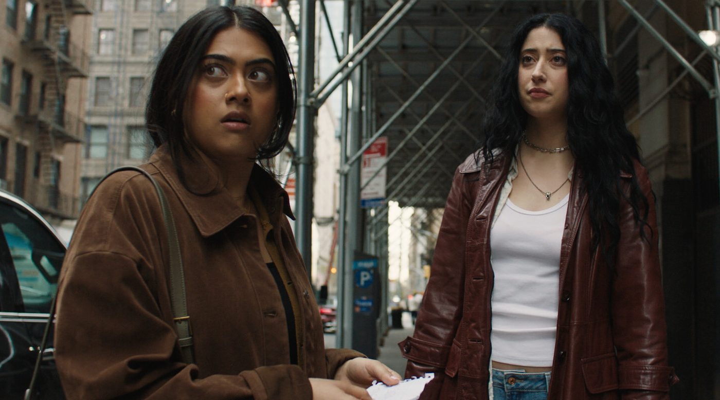Two women stand on a New York sidewalk under construction scaffolding, both looking up at something off to one side.