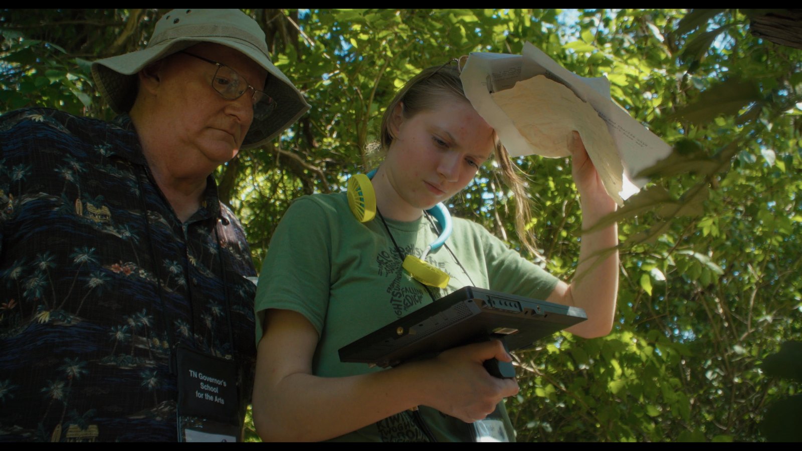 An instructor in a sun hat and a student looking at a tablet outdoors, a script in her other hand