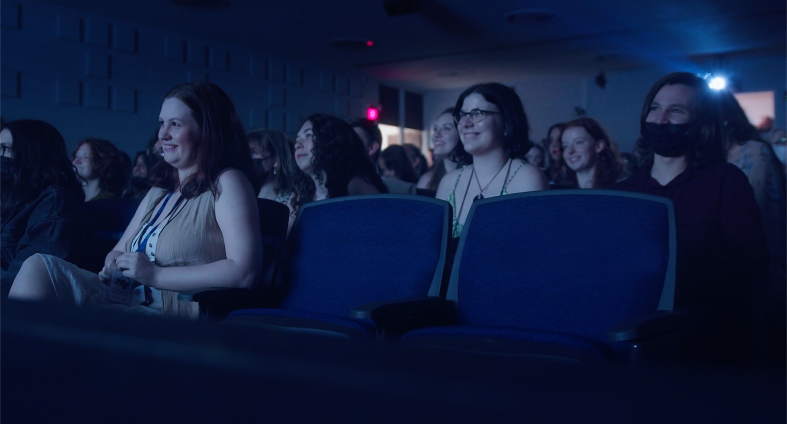 An audience in a dark theater lit blue by the screen, smiling