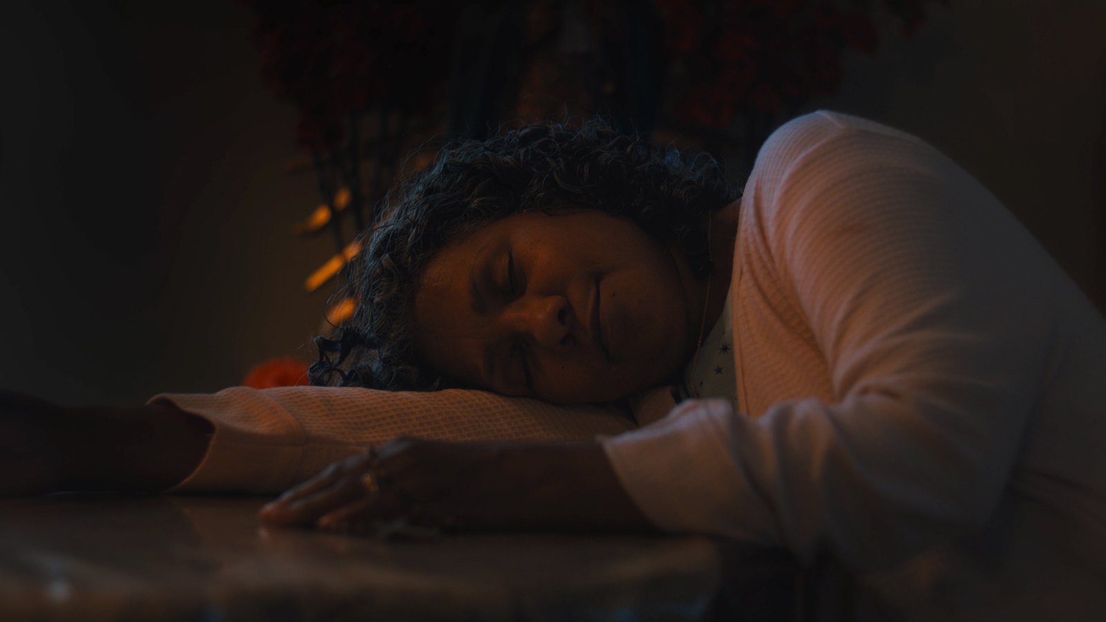 Luz asleep with her head on her arm at a table, in low warm light