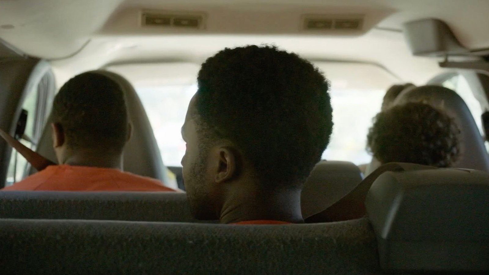 The inside of a van seen from the back seat: young men from behind, two of them in orange