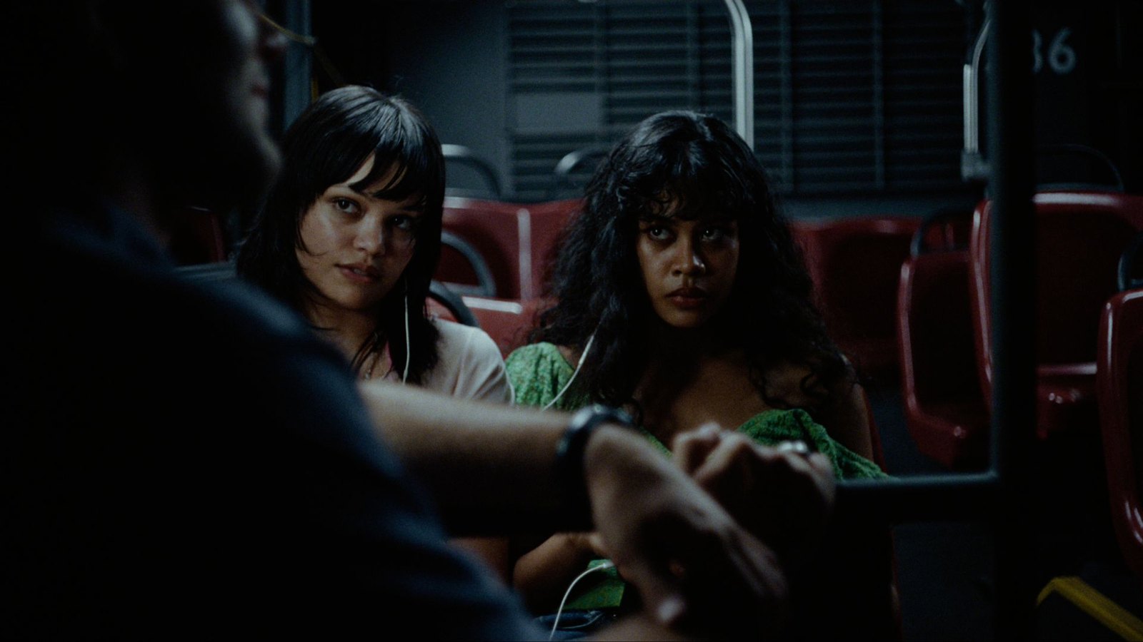 Two teenage girls share a pair of earbuds on a night bus with red seats; in the foreground, out of focus, a man's arm and wristwatch