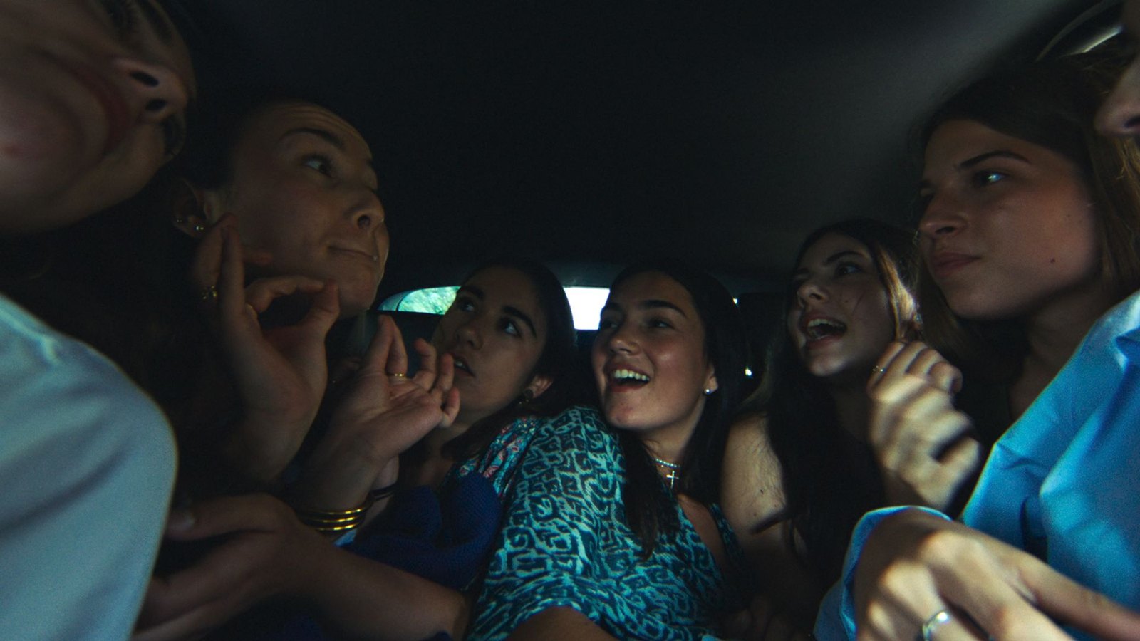 Six young women crowded into a car, laughing and talking, seen from the front seat