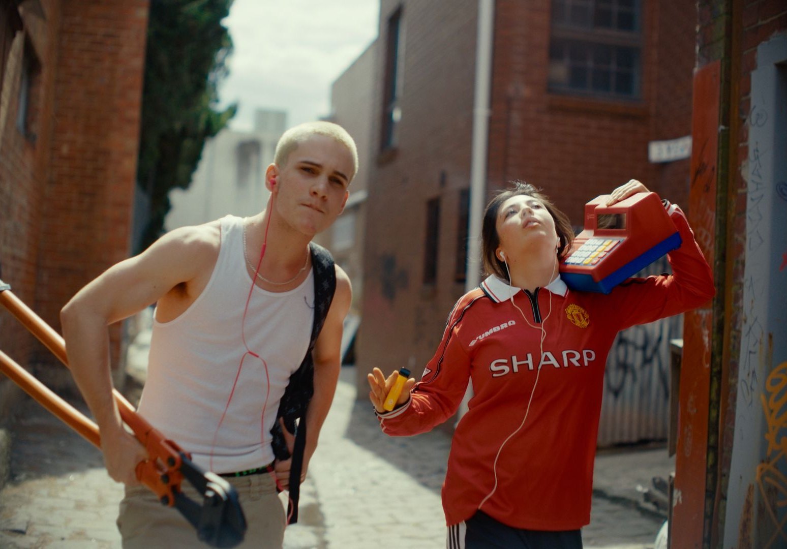 Two friends in a brick laneway: a young man with bleached hair holding long orange bolt cutters, and a young woman in a red football shirt with a child's toy cash register on her shoulder