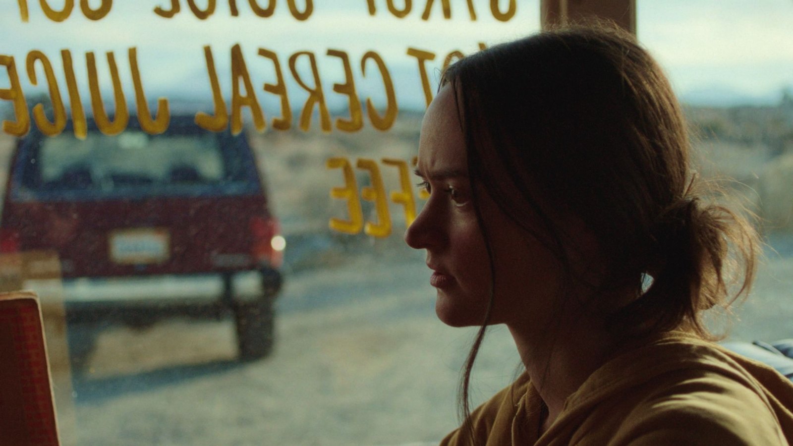 A young woman in profile at a diner window, frowning; outside, a dark red Jeep is parked in the desert