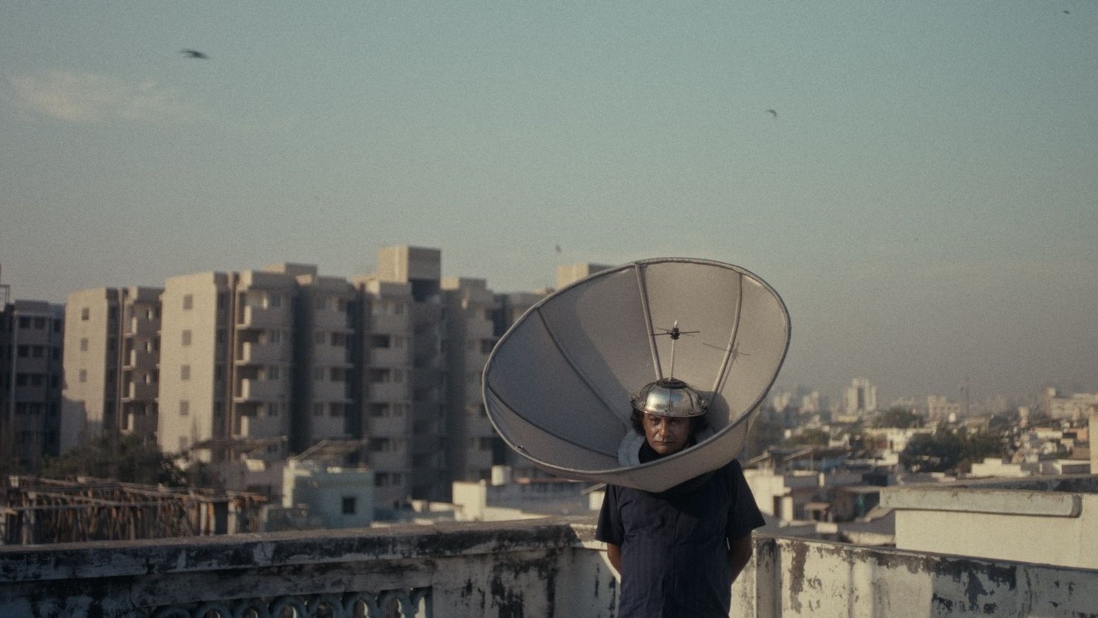 A man stands on a city rooftop with a large satellite dish around his head and a steel bowl worn like a helmet, apartment blocks behind him