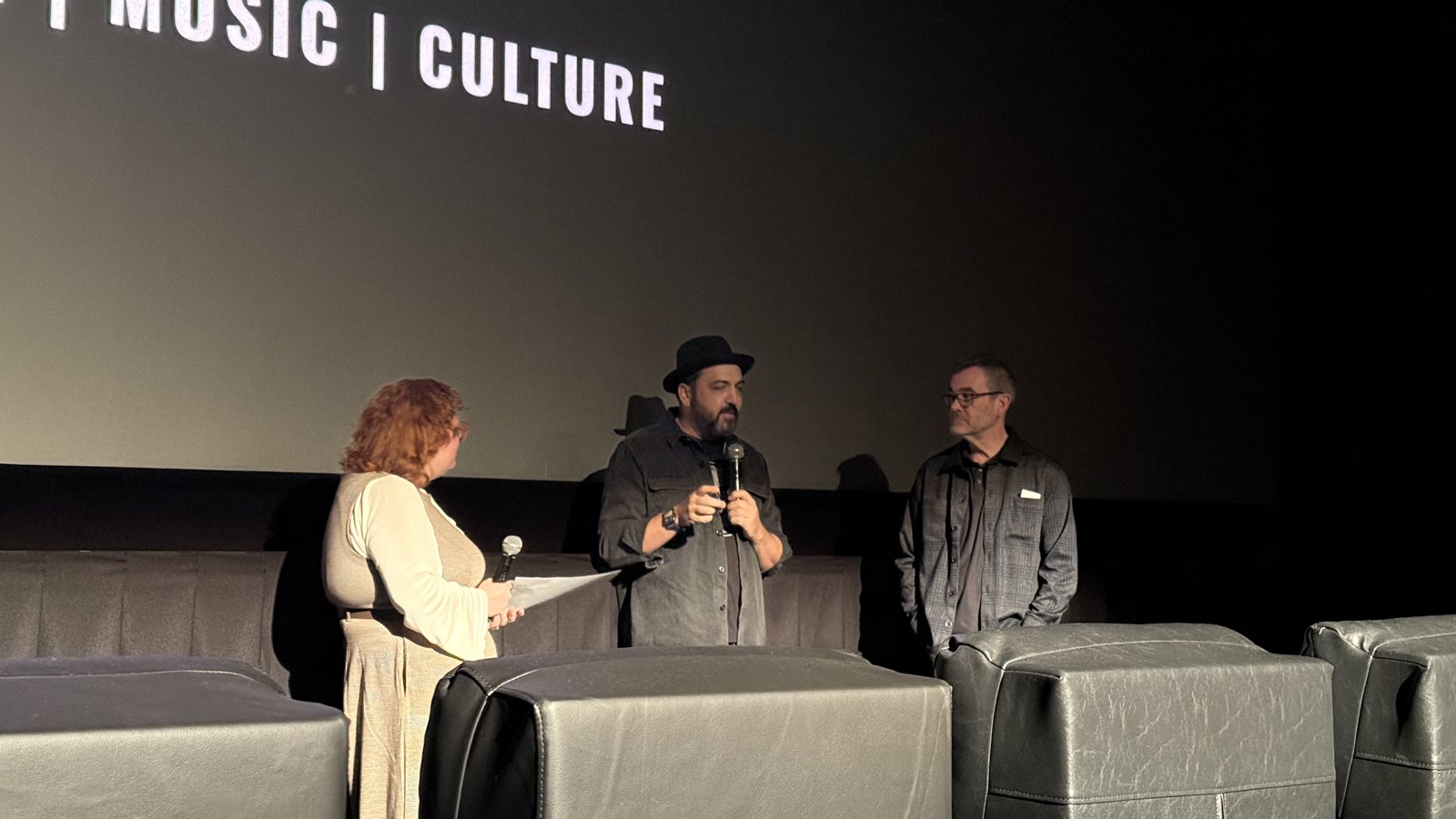 Three people stand at the front of a cinema under the Nashville Film Festival logo; the man in the middle, in a hat, holds a microphone.