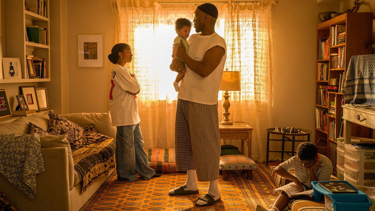 Latif holding his baby in a sunlit living room while his daughter watches and his son reads on the floor