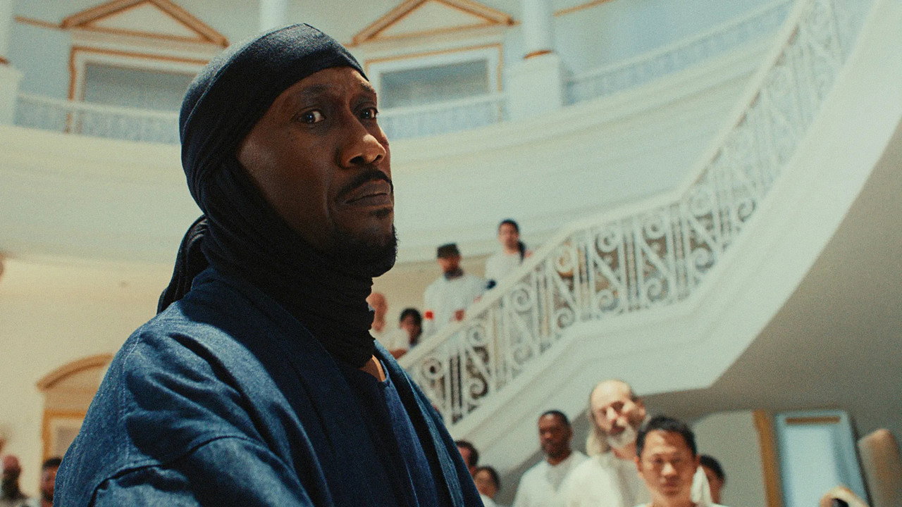Latif in a dark head wrap in a white mosque, men gathered on the staircase behind him