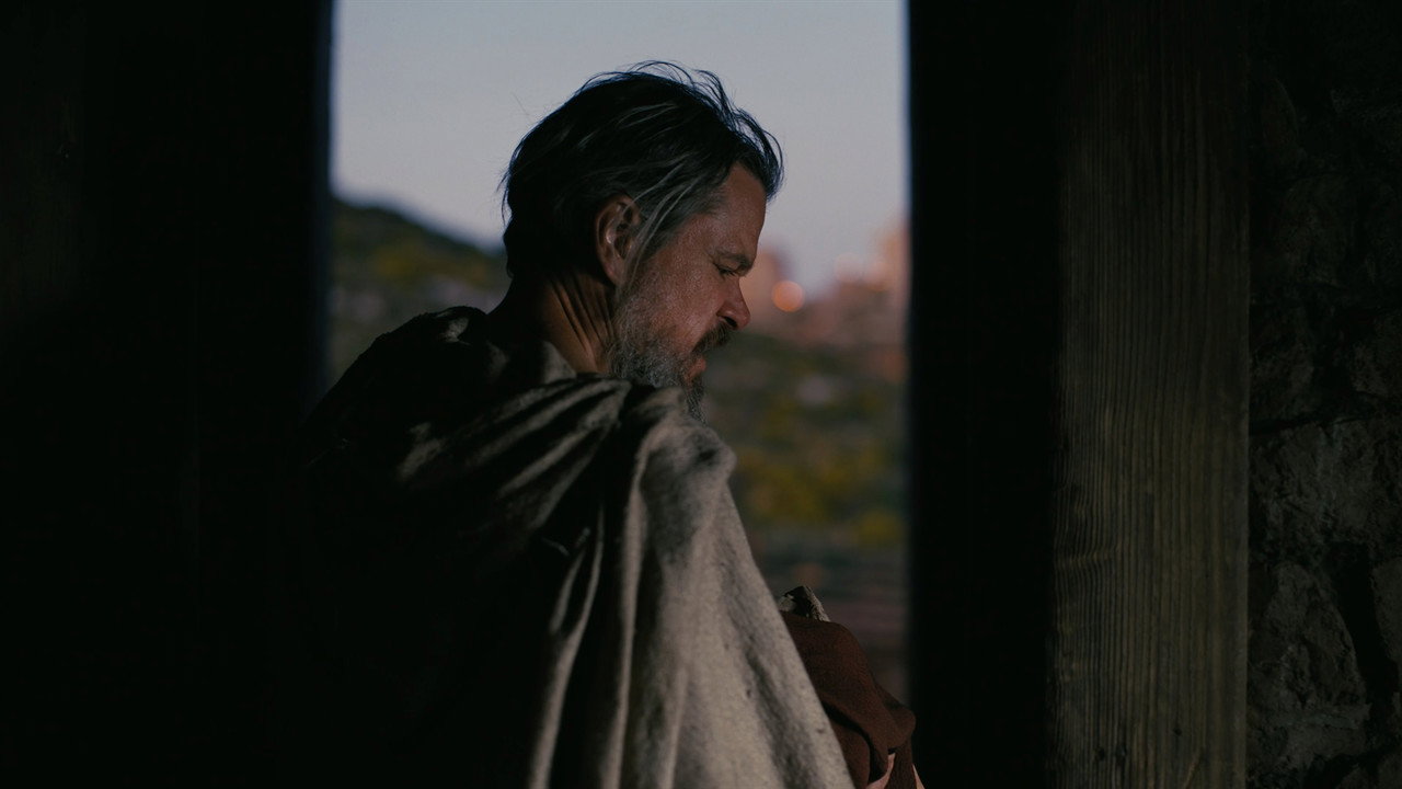 A cloaked Odysseus stands at an interior threshold at dusk, from Christopher Nolan's The Odyssey